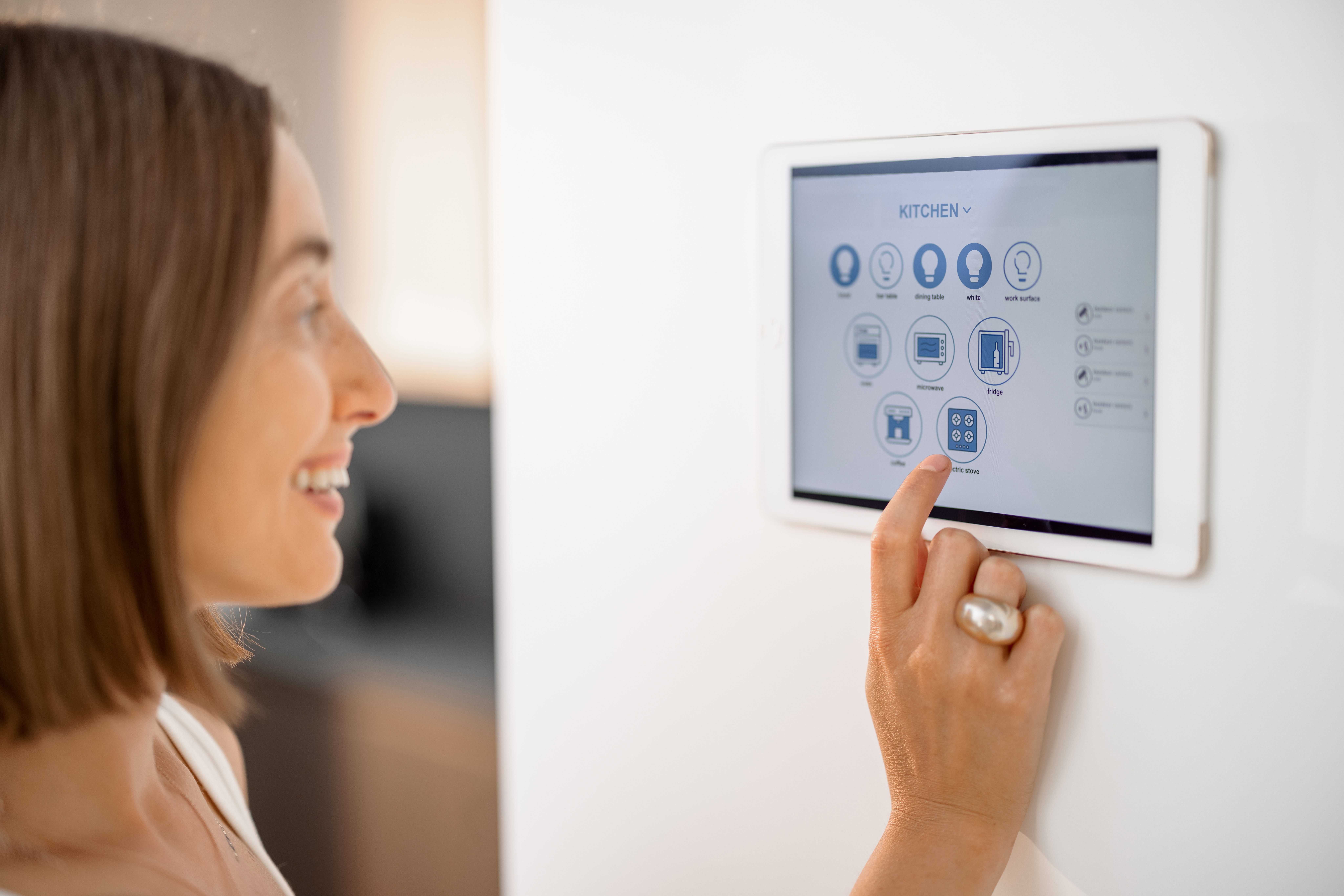 Happy woman controlling smart devices using control panel mounted on the wall at kitchen. Digital tablet with running mobile application. Smart home concept